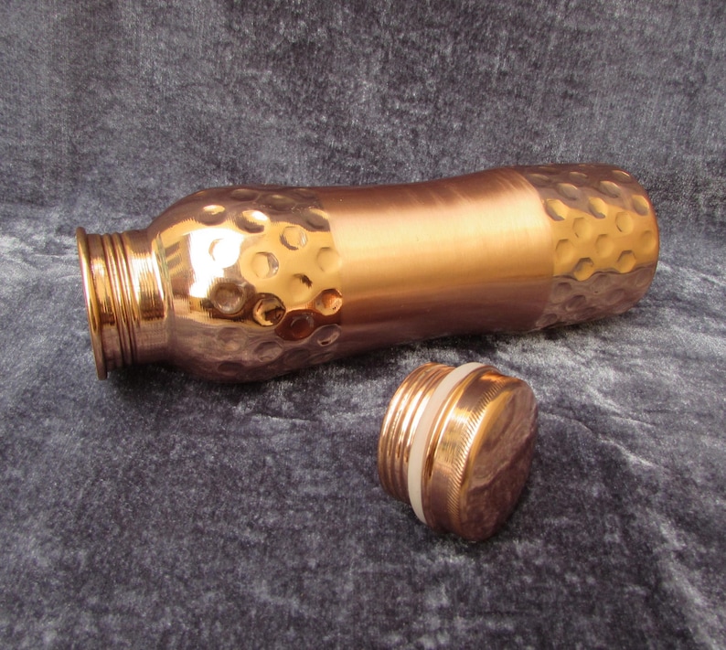 May include: A copper water bottle with a hammered texture on the top and bottom sections. The bottle is lying on its side with the cap unscrewed and next to it. The bottle has a smooth, polished finish in the middle section.