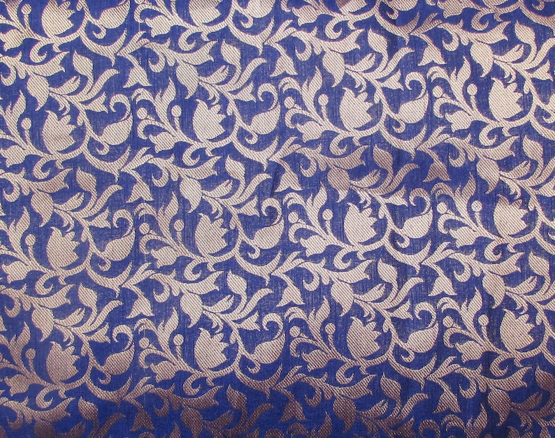 May include: Close-up of a blue fabric with a repeating floral pattern in a lighter, metallic gold color. The design features stylized flowers and leaves, creating a textured, ornate appearance. The fabric has a slight sheen.