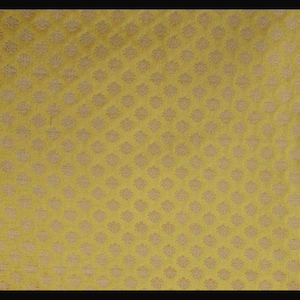 May include: A mustard yellow fabric featuring a repeating pattern of small, light-colored floral motifs arranged in a grid. The textile has a visible woven texture with a consistent, dense arrangement of these symmetrical, stylized flower shapes.