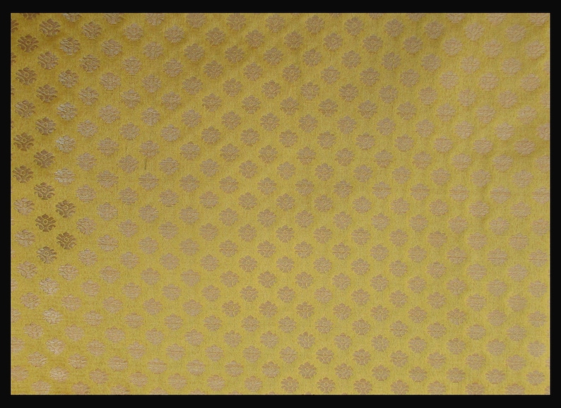 May include: A mustard yellow fabric featuring a repeating pattern of small, light-colored floral motifs arranged in a grid. The textile has a visible woven texture with a consistent, dense arrangement of these symmetrical, stylized flower shapes.