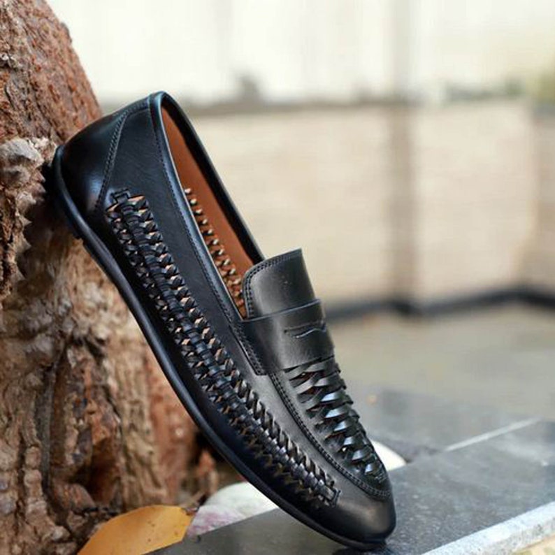 May include: A black slip-on loafer featuring a woven leather side panel and a smooth leather vamp. The shoe has a low profile, a rounded toe, and a tan interior lining. The design incorporates a decorative horizontal strap across the top of the foot.