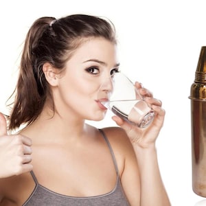 May include: A woman drinks water from a glass, giving a thumbs-up gesture. A tall, metallic cocktail shaker with a gold-colored top sits to the right. The background is plain white.