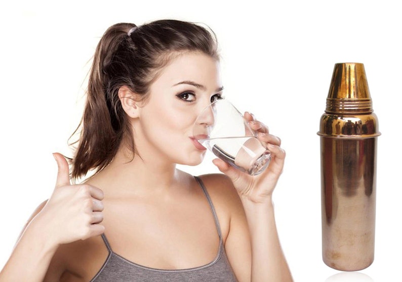 May include: A woman drinks water from a glass, giving a thumbs-up gesture. A tall, metallic cocktail shaker with a gold-colored top sits to the right. The background is plain white.