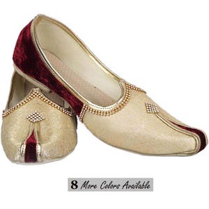 May include: A pair of slip-on shoes featuring a gold-toned fabric with a subtle embossed pattern, accented by deep maroon velvet panels. The shoes are decorated with gold piping and small, clear crystal embellishments. Text reads 8 More Colors Available.