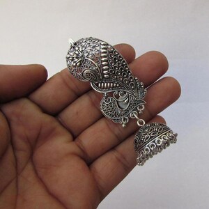 May include: Silver earring with an ornate paisley design and a bell-shaped drop. The earring features detailed filigree work and a small, round bell at the bottom. The design includes floral and peacock-inspired elements.