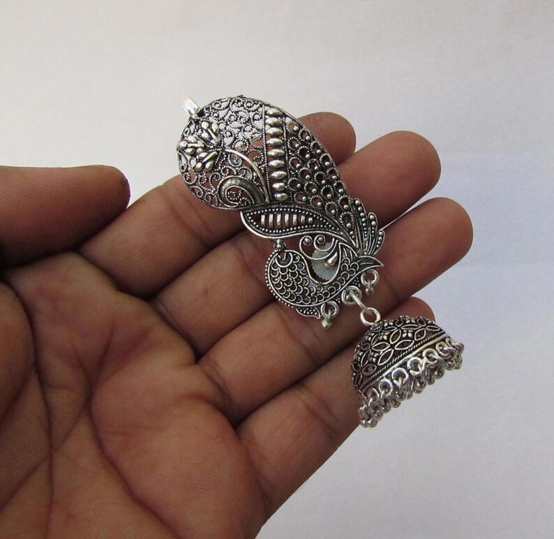 May include: Silver earring with an ornate paisley design and a bell-shaped drop. The earring features detailed filigree work and a small, round bell at the bottom. The design includes floral and peacock-inspired elements.