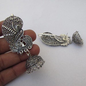 May include: Silver-coloured peacock-shaped earrings. The earrings have detailed filigree patterns and a dangling bell-shaped charm. The design includes a peacock motif with a detailed feather pattern. The earrings are displayed on a white surface.