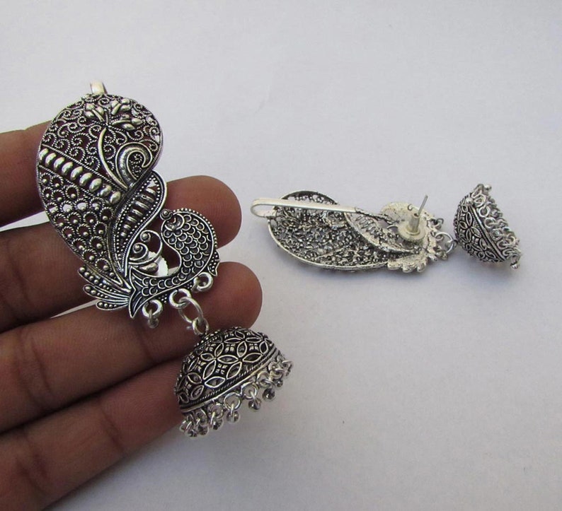 May include: Silver-coloured peacock-shaped earrings. The earrings have detailed filigree patterns and a dangling bell-shaped charm. The design includes a peacock motif with a detailed feather pattern. The earrings are displayed on a white surface.