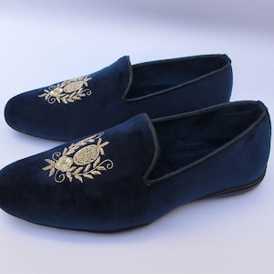 Pode incluir: Um par de mocassins de veludo azul-marinho com um motivo floral e oval bordado a fio dourado no peito do p&eacute;. Os sapatos sem atacadores t&ecirc;m biqueira arredondada, perfil baixo, um debrum preto ao longo da abertura e uma sola preta plana.