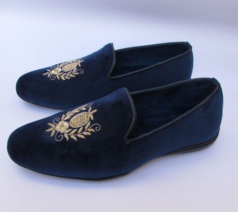 Pode incluir: Um par de mocassins de veludo azul-marinho com um motivo floral e oval bordado a fio dourado no peito do p&eacute;. Os sapatos sem atacadores t&ecirc;m biqueira arredondada, perfil baixo, um debrum preto ao longo da abertura e uma sola preta plana.