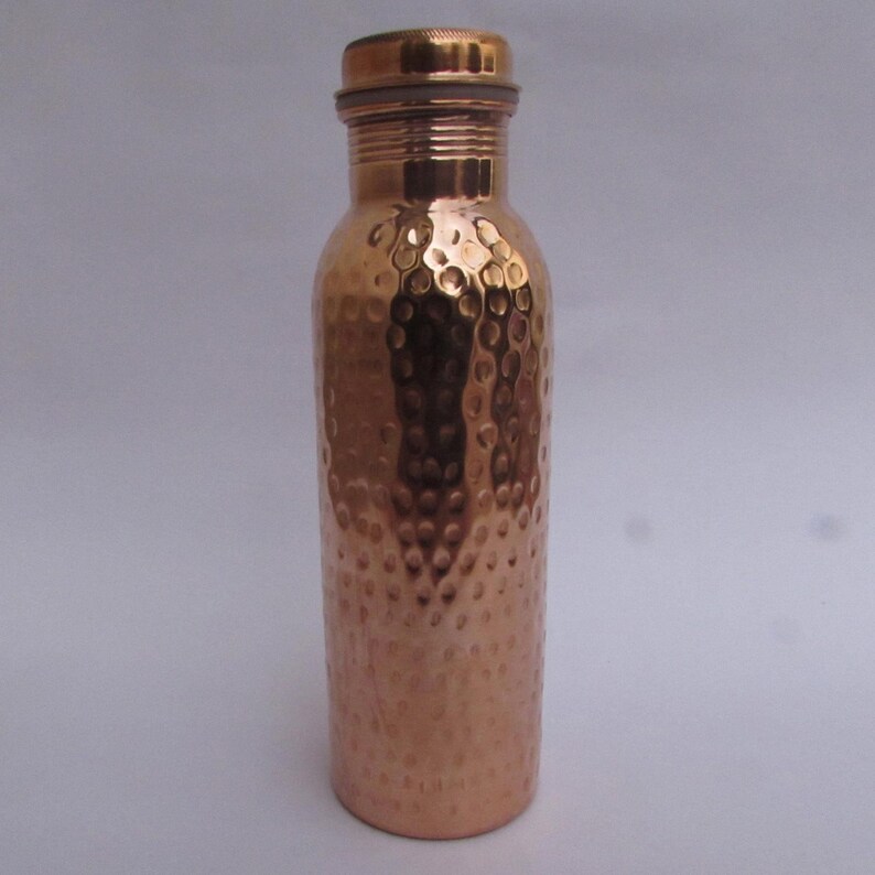 May include: A cylindrical copper water bottle featuring a distinct hammered texture across its body. The bottle has a smooth, narrow neck and a matching metallic screw-top lid with a textured rim, showcasing a polished, reflective reddish-brown finish.