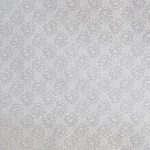 May include: A close-up of a white fabric with a repeating pattern of stylized floral and paisley designs. The pattern is slightly raised, creating a subtle texture. The background is a solid white.
