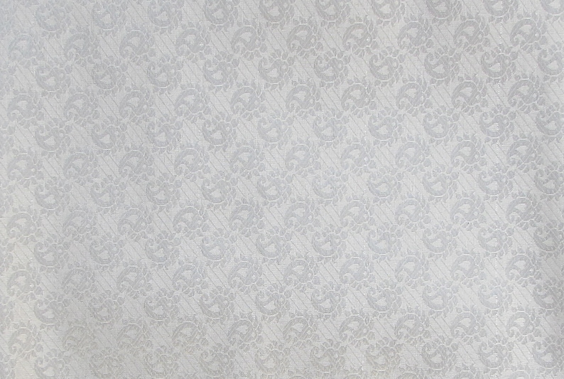May include: A close-up of a white fabric with a repeating pattern of stylized floral and paisley designs. The pattern is slightly raised, creating a subtle texture. The background is a solid white.