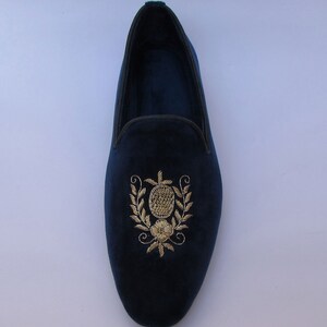 Pode incluir: Um mocassim de veludo azul-marinho com bordado em fio dourado no peito do p&eacute;. O bordado retrata uma forma oval central rodeada por ramos de folhas e uma pequena flor na base. O sapato tem um acabamento escuro ao longo da abertura.