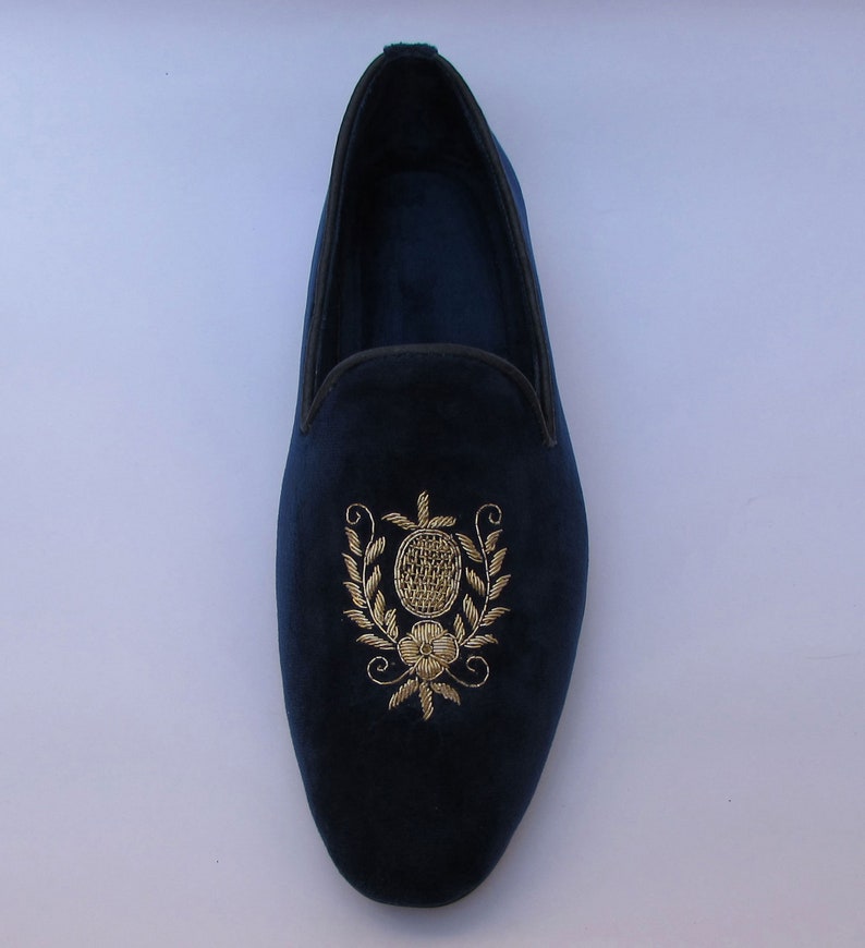 Pode incluir: Um mocassim de veludo azul-marinho com bordado em fio dourado no peito do p&eacute;. O bordado retrata uma forma oval central rodeada por ramos de folhas e uma pequena flor na base. O sapato tem um acabamento escuro ao longo da abertura.