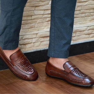 May include: A pair of brown leather loafers featuring a woven texture across the vamp and sides. The shoes have a classic penny loafer strap detail across the top and a smooth, polished finish on the toe and heel sections with a dark sole.