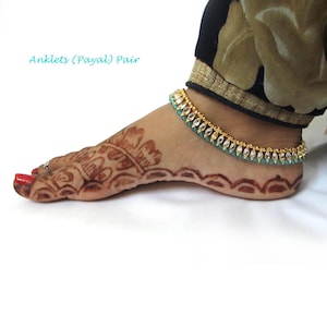 May include: A pair of gold-toned anklets featuring a row of clear, teardrop-shaped stones accented by small, round teal beads. The anklets are worn around the ankle, which displays intricate brown henna patterns and a single silver-toned toe ring.