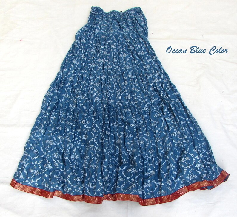 May include: A long, crinkled skirt in an ocean blue color featuring a white floral vine pattern throughout. The skirt has an elasticized waistband and a wide, contrasting dark red border at the hem with a subtle gold-toned dotted detail.