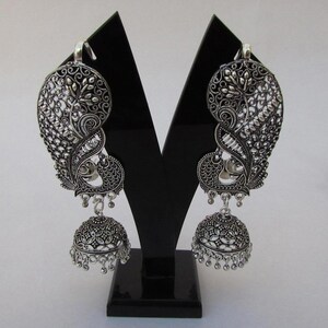 May include: Silver-coloured peacock-shaped earrings with detailed filigree patterns. The earrings have a large, fan-shaped design with a peacock motif, and a dangling bell-shaped charm. The earrings are displayed on a black stand against a white background.