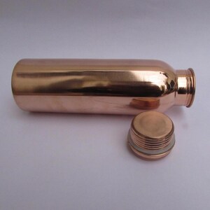 May include: A copper water bottle with a screw-on lid. The bottle is cylindrical with a polished finish, reflecting the light. The lid is detached, showing the threaded opening. The overall design is simple and functional.
