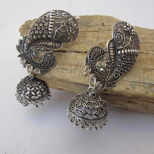 May include: A pair of silver earrings with a peacock design. The earrings have a detailed, openwork bell shape suspended from a peacock-shaped top. The peacock design includes intricate feather patterns and a curved shape.