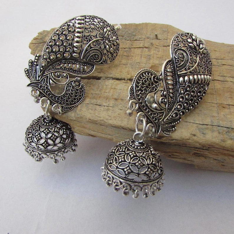 May include: A pair of silver earrings with a peacock design. The earrings have a detailed, openwork bell shape suspended from a peacock-shaped top. The peacock design includes intricate feather patterns and a curved shape.