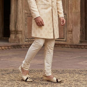 May include: A cream-colored sherwani jacket with a subtle gold floral pattern, featuring a high collar and front buttons, paired with matching cream-colored slim-fit trousers and pointed-toe slip-on shoes with dark brown and gold accents.
