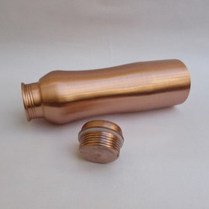 May include: A copper-colored water bottle with a matching screw-on cap. The bottle has a curved, ergonomic design and a threaded neck. The cap is detached, showing the threaded interior and a white seal. The bottle is made of metal.