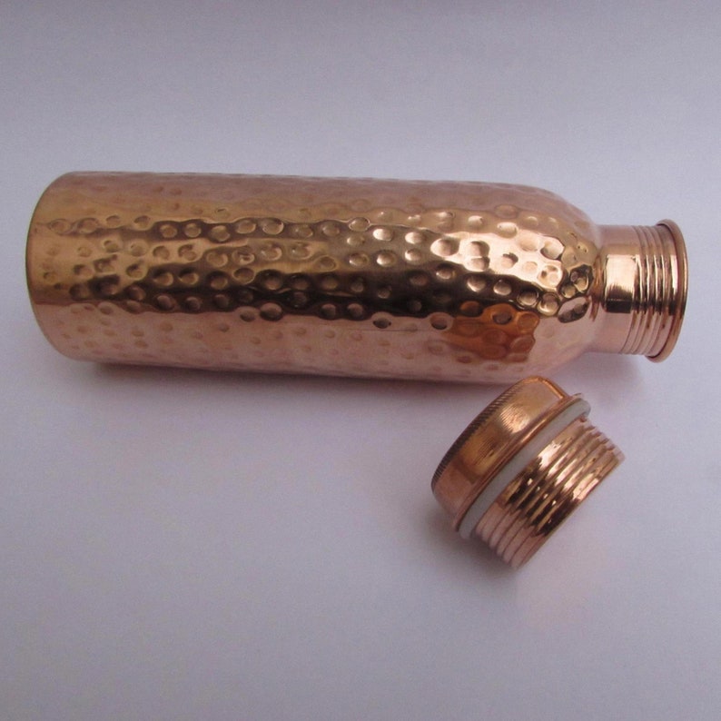 May include: A copper water bottle with a hammered texture and a screw-on cap. The bottle is cylindrical and has a shiny, metallic finish. The cap is detached, revealing the threaded opening. The bottle is approximately 10 inches tall.