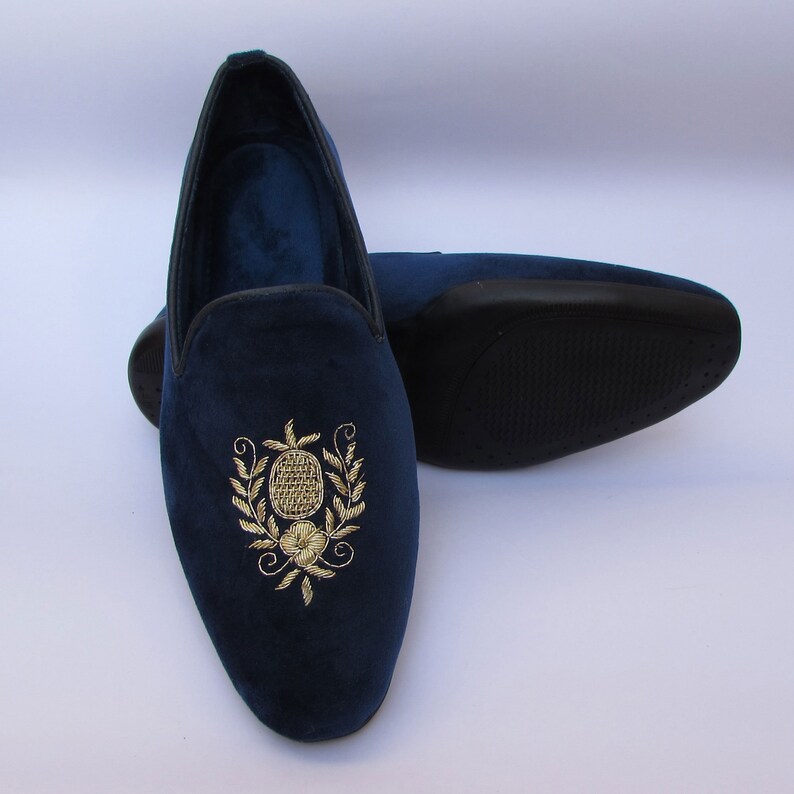 Pode incluir: Um par de mocassins de veludo azul-marinho com um bordado dourado de abacaxi e motivos florais no peito do p&eacute;. Os sapatos t&ecirc;m um rebordo escuro &agrave; volta da abertura e uma sola preta texturada no segundo sapato posicionado atr&aacute;s.