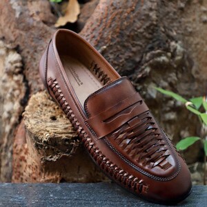 May include: A brown leather loafer featuring a woven design on the upper and sides. The shoe has a classic penny loafer strap across the vamp. The interior insole is printed with the text 'AimHeeShoes' and 'aimheeshoes.com' in black ink.