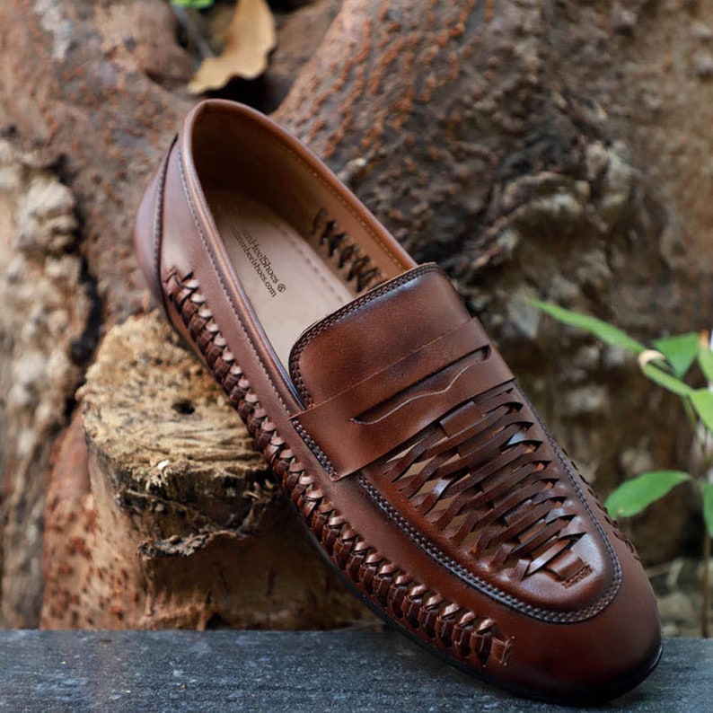 May include: A brown leather loafer featuring a woven design on the upper and sides. The shoe has a classic penny loafer strap across the vamp. The interior insole is printed with the text 'AimHeeShoes' and 'aimheeshoes.com' in black ink.