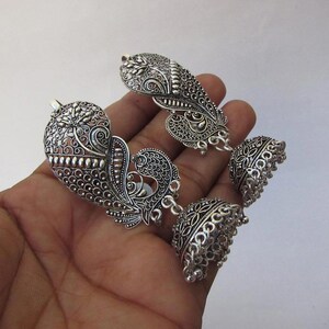 May include: Silver earrings with detailed filigree and peacock motifs. The earrings have a large, heart-shaped top and a bell-shaped bottom, displaying intricate patterns and textures. The earrings are presented against a plain background.