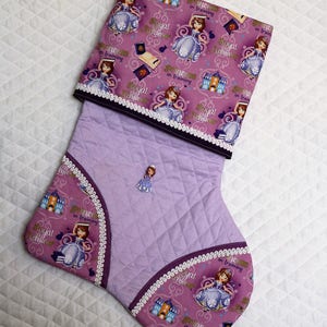 May include: Purple and pink quilted stocking with a white lace trim. The stocking features a repeating pattern of a princess in training, a castle, and the words "Royal Achievers".