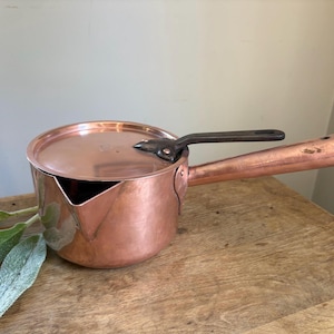 May include: A polished copper saucepan with a matching lid and a long, straight copper handle. The pan features a distinct pouring spout on the side and a dark, metallic handle attachment secured with rivets to the lid.