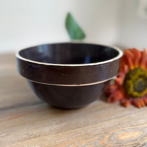 May include: A dark brown ceramic bowl with a white rim. The bowl is sitting on a wooden surface. A decorative orange flower is in the background.