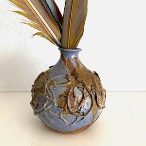 May include: A round ceramic vase with a narrow neck, featuring a mottled blue and brown glaze with raised, swirling clay textures on the surface. Three large, long feathers in shades of yellow, brown, and teal are placed inside the vase opening.