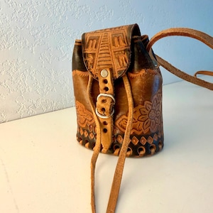 May include: A brown leather drawstring bag featuring intricate embossed floral and geometric patterns. The bag has a flap closure secured with a metal buckle and a long leather shoulder strap. The base is detailed with a series of cutouts.