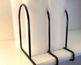 May include: A black metal shelf divider is holding a white foam block on a white shelf. The divider has a curved top and two vertical supports. The shelf is mounted on a white wall.