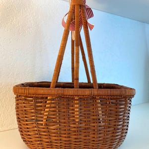 May include: A woven brown wicker basket with a rectangular base and a tall handle. The handle is attached to the basket with four vertical supports. A red and white striped ribbon is tied to the top of the handle.