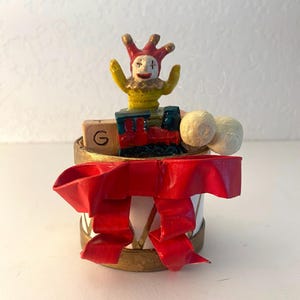 May include: A decorative Christmas ornament featuring a white cylinder with a gold rim and a large red bow. The ornament has a jester figure with a red and yellow hat, a small red train, and two white balls.
