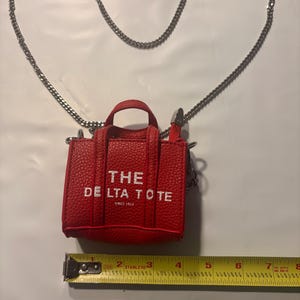 Könnte beinhalten: Eine kleine, rote Tragetasche mit einem silbernen Kettenriemen. Die Tasche hat eine strukturierte Oberfläche und die Worte "THE DELTA TOTE SINCE 1915" sind in Weiß aufgedruckt. Die Tasche ist etwa 10 cm breit.