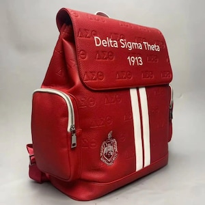 Peut inclure: Sac à dos rouge avec des détails blancs et le texte "Delta Sigma Theta 1913". Le sac présente une surface texturée, des rayures verticales blanches et un emblème argenté. Il possède une poche zippée et une fermeture à rabat.