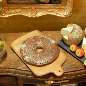 May include: A brown, round, cake-like dessert with a hole in the center, sitting on a wooden cutting board. The dessert is surrounded by various cheeses, fruits, and other snacks.