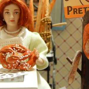 May include: A woman in a white lace top holds a pretzel with salt. The pretzel is brown and has a white glaze. The background is a wooden shelf with other pretzels.