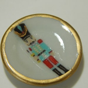 May include: A small, round, white porcelain plate with a gold rim. The plate features a hand-painted image of a toy soldier in a blue and red uniform.