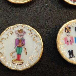 May include: A white porcelain plate with a gold rim and a hand-painted image of a mouse dressed as a nutcracker. The plate has a gold-painted border with a repeating pattern of dots and swirls.