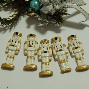 May include: Five white and gold nutcracker figurines with blue eyes. The nutcrackers are standing upright on a white surface.