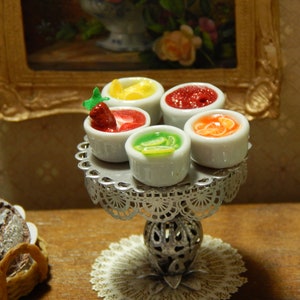 May include: A silver tiered stand with five small white bowls filled with different colored fruit preserves. The bowls are arranged in a circle on the stand. The stand is on a white lace doily.
