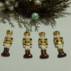 May include: Four miniature nutcracker figurines with gold and white paint, blue eyes, and black accents. The figurines are standing on a white surface.